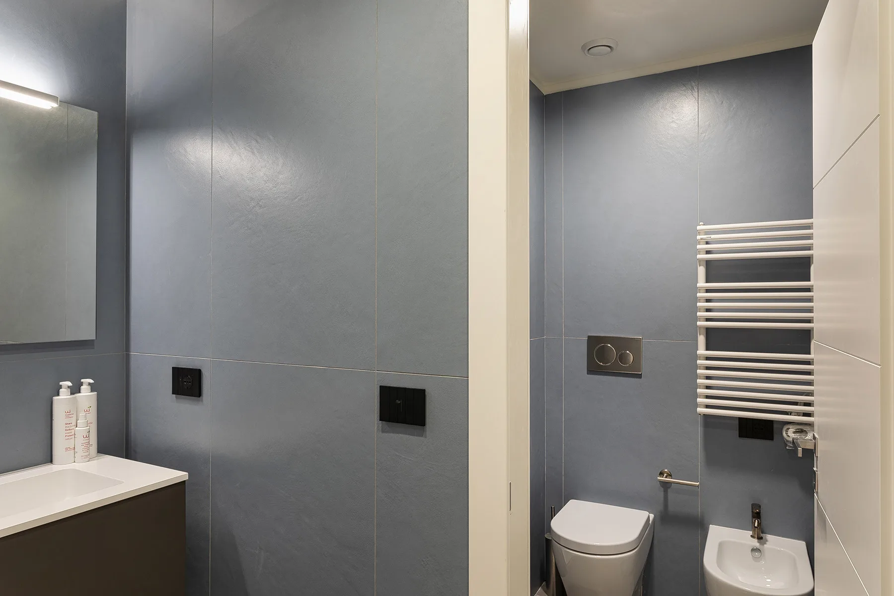 A modern bathroom with gray tiled walls, a toilet, and a bidet.