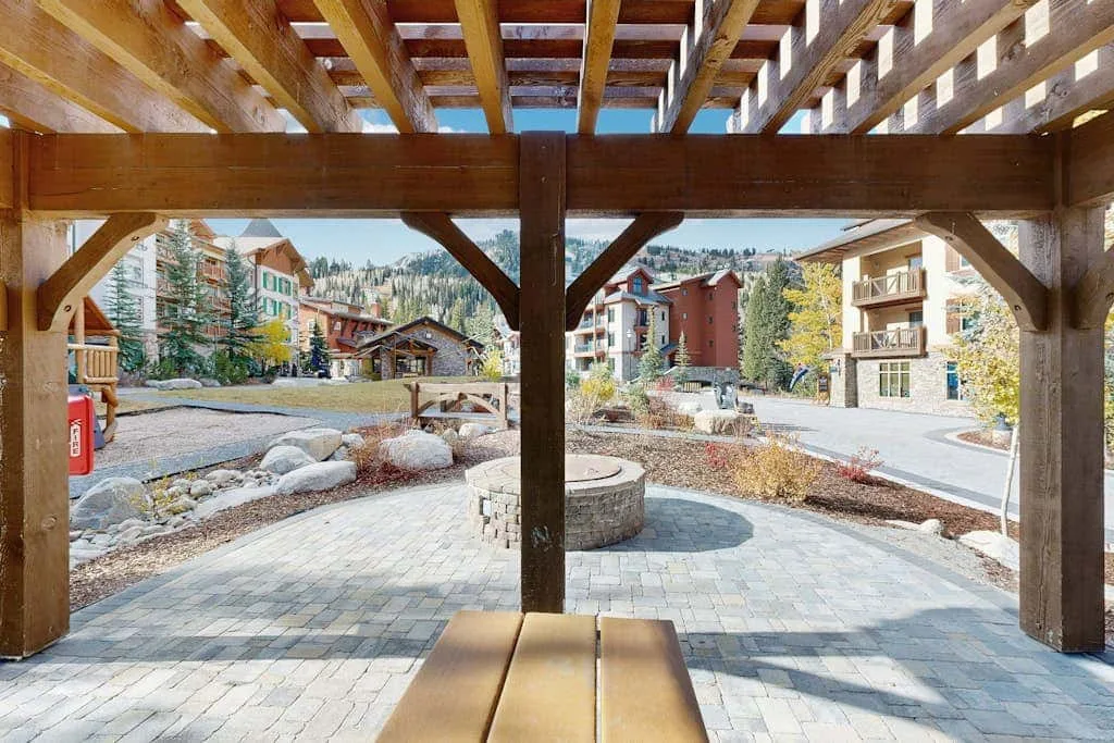 Un patio exterior pavimentado bajo una pérgola de madera da a edificios cercanos.