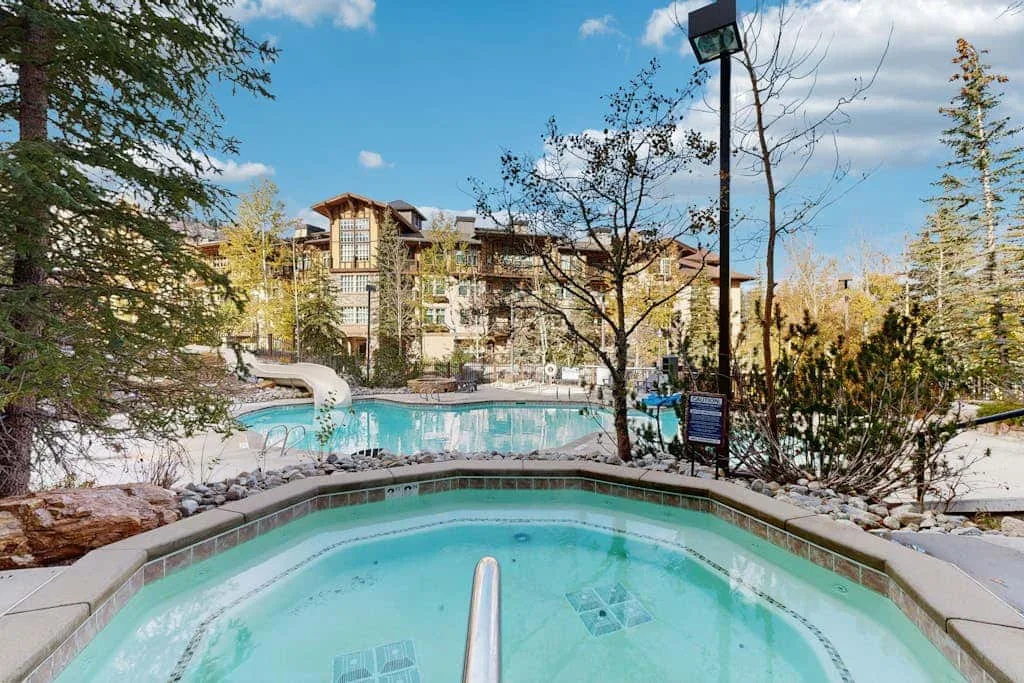 Una piscina exterior y una bañera de hidromasaje están entre árboles, con un edificio de varias plantas al fondo.