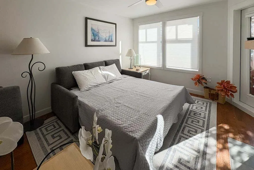 Un dormitorio con una cama, una obra enmarcada y ventanas luminosas.