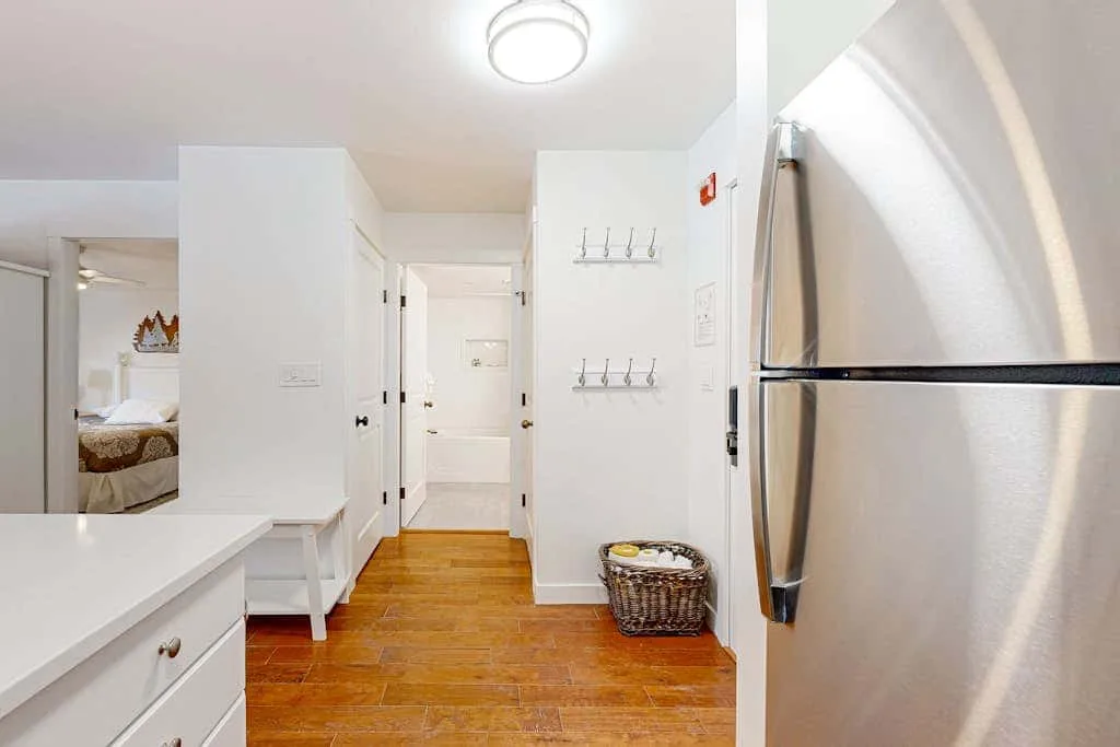 Un pasillo luminoso con piso de madera, ganchos en la pared y un refrigerador.