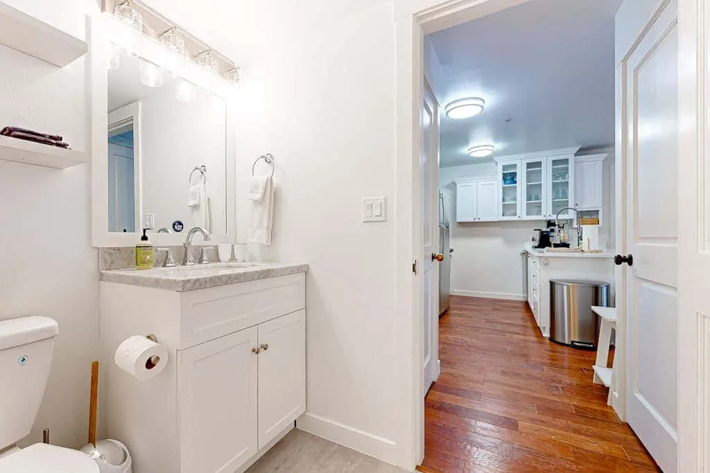Un tocador de baño da a una cocina con gabinetes blancos y piso de madera.