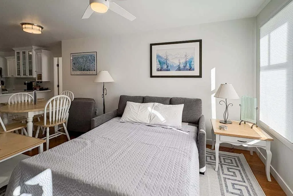 Un dormitorio con una cama, asientos grises y una ventana luminosa.