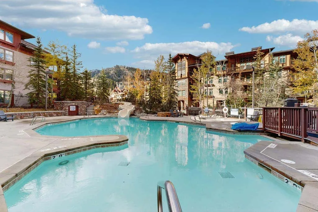 An outdoor swimming pool sits among multi-story buildings and trees.