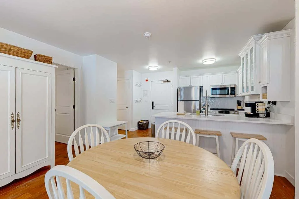 Una cocina y comedor luminosos con gabinetes blancos y una mesa redonda de madera.