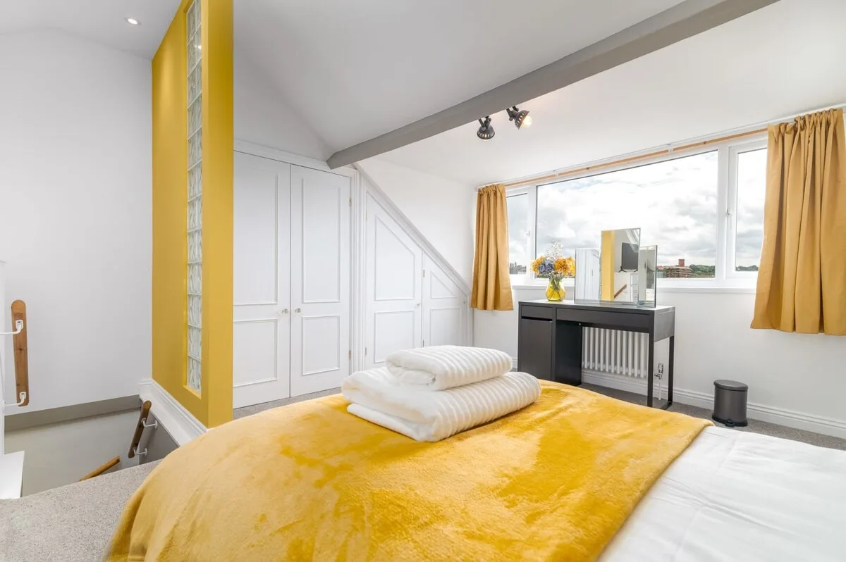 A bright bedroom with a bed covered by a mustard-yellow blanket and a window.