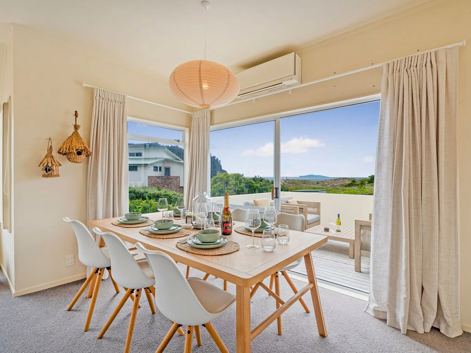 Un comedor con una mesa de madera y seis sillas blancas junto a grandes ventanas y una puerta corredera que da a una zona de estar al aire libre en Sans Souchi - Whangamata Holiday Home, en Whangamata