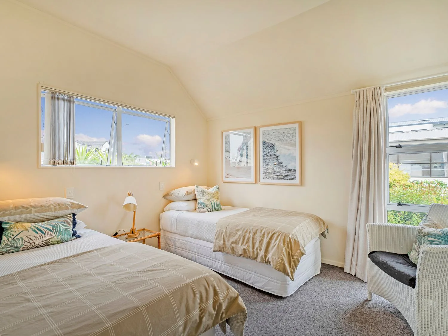 Un dormitorio con dos camas individuales, una mesita de noche, una lámpara, dos cuadros enmarcados y un sillón de mimbre en Sans Souchi - Whangamata Holiday Home, en Whangamata