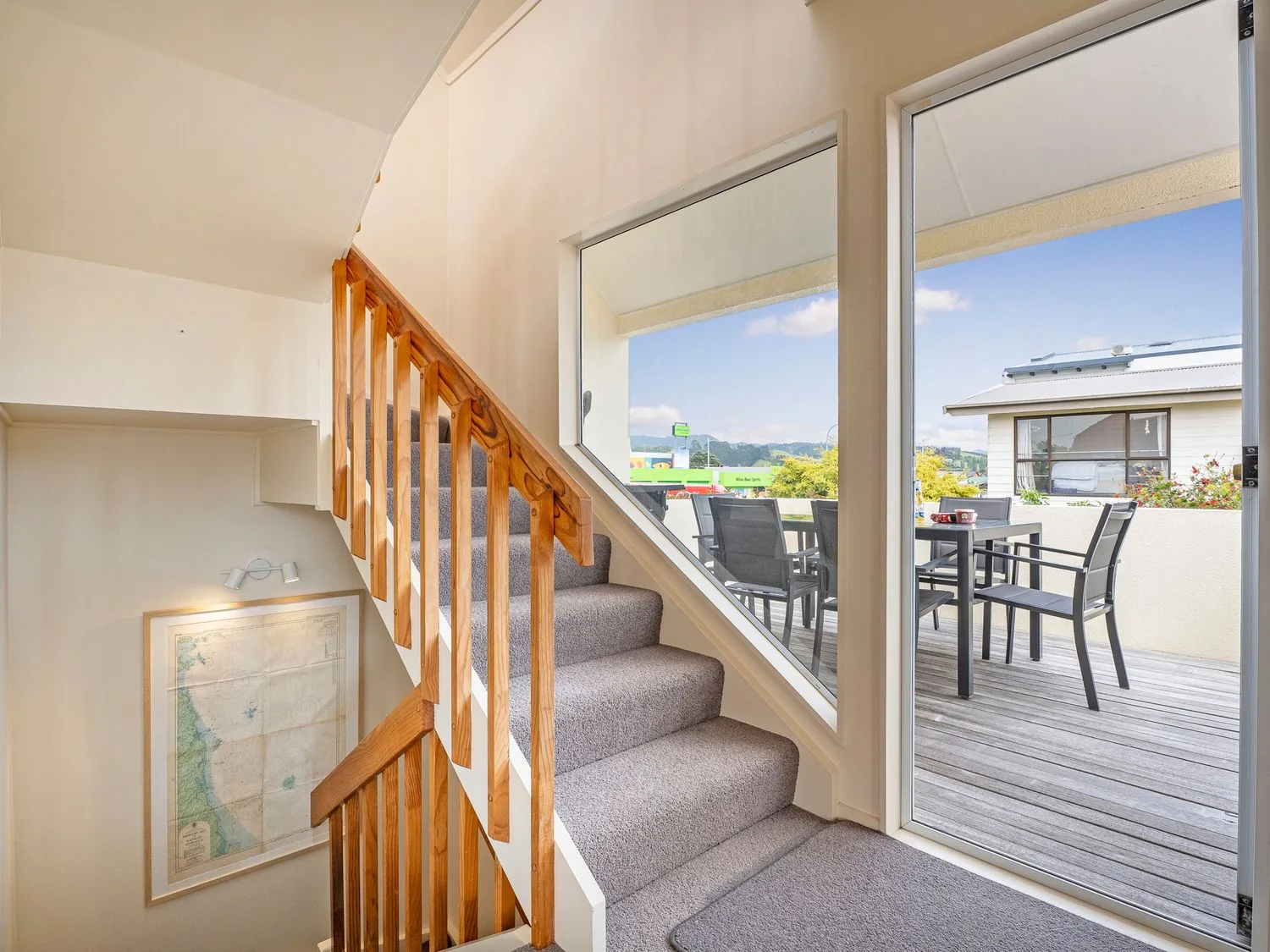 Una escalera con barandilla de madera junto a una gran ventana con vistas a una terraza de madera con mesa y sillas de exterior en Sans Souchi - Whangamata Holiday Home, en Whangamata