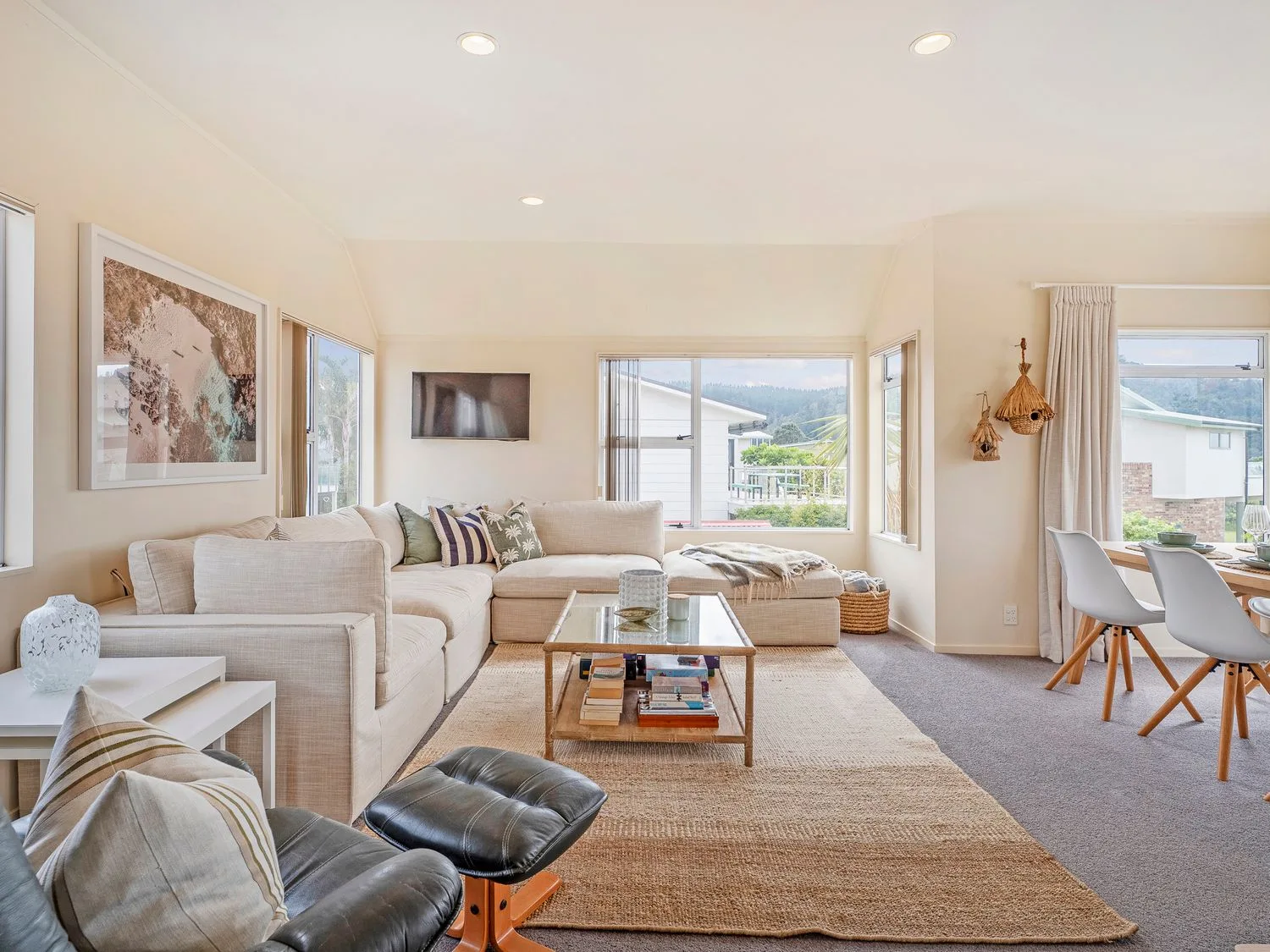 A living room with a beige sectional sofa a glass coffee table a leather chair and ottoman and a dining area with white chairs at Sans Souchi - Whangamata Holiday Home in Whangamata
