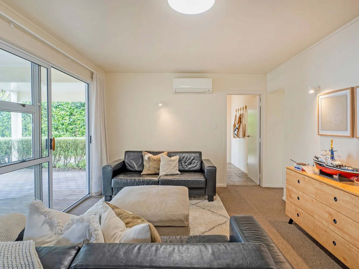 Una sala de estar con asientos oscuros, un reposapiés beige y puertas corredizas de vidrio que dan a un patio.