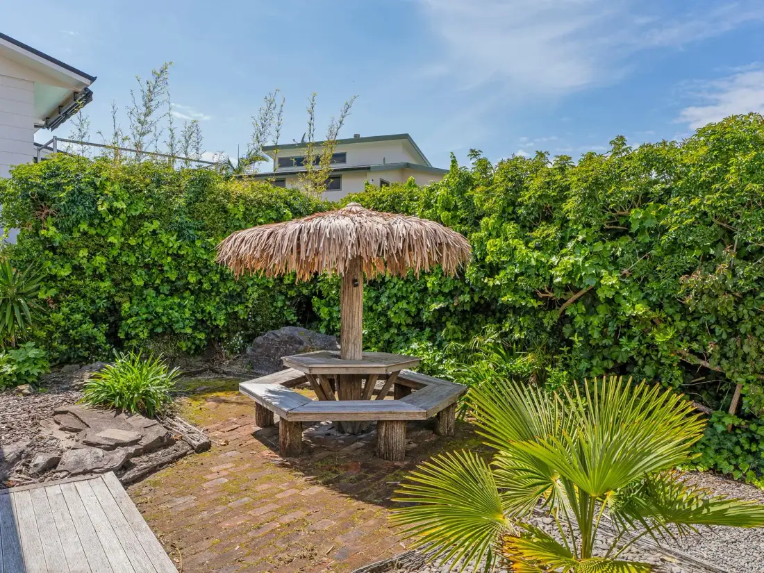 Una zona de jardín con un conjunto de banco y mesa de madera bajo una sombrilla de paja, rodeado de plantas y vegetación, en Sans Souchi - Whangamata Holiday Home, en Whangamata
