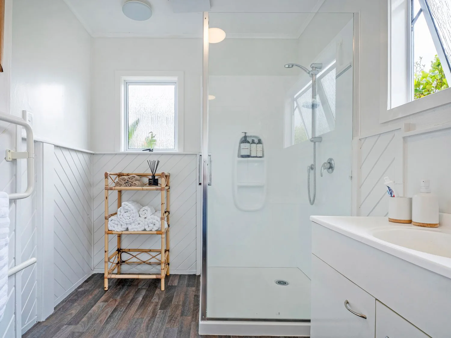 Un baño luminoso con ducha de cristal, toallero de madera y mueble lavabo blanco.