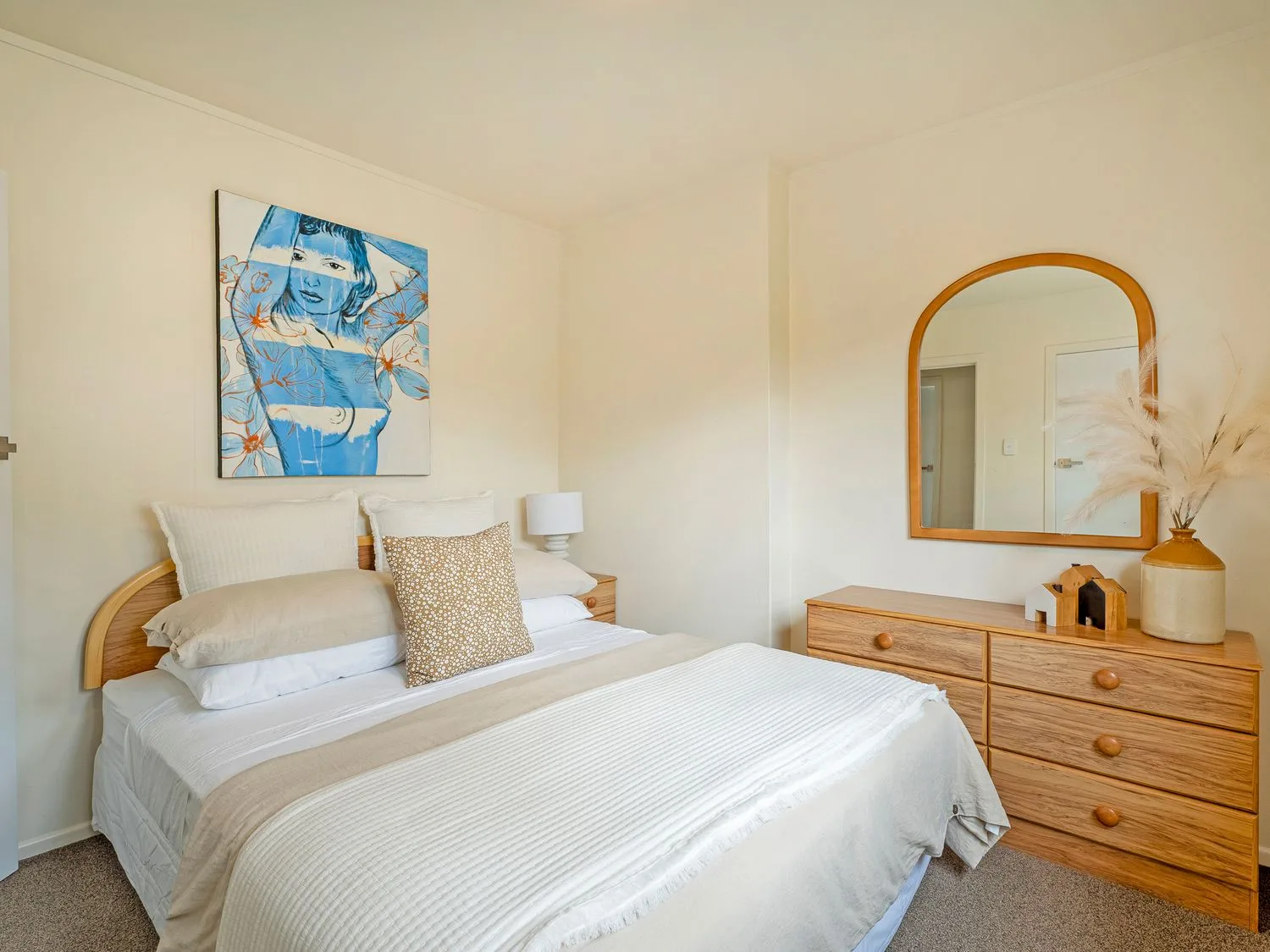 Un dormitorio con una cama, almohadas, una lámpara de noche, una cómoda de madera con espejo y objetos decorativos en Sans Souchi - Whangamata Holiday Home, en Whangamata