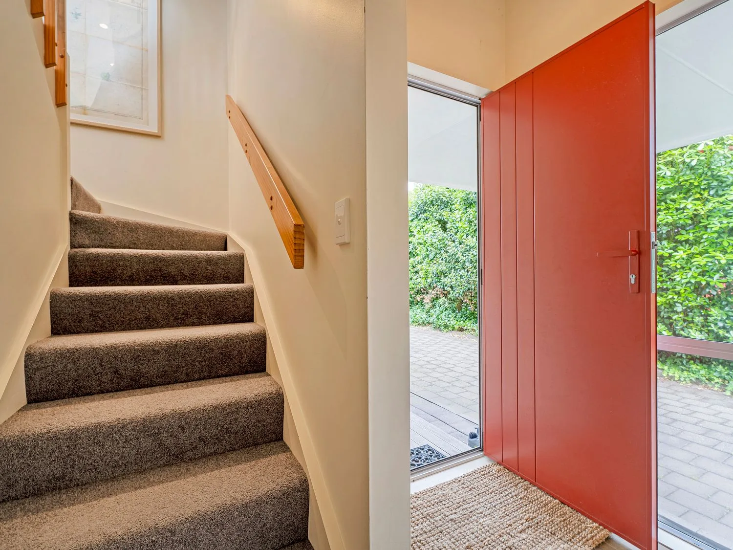Una escalera enmoquetada junto a una puerta de entrada roja abierta con un felpudo en Sans Souchi - Whangamata Holiday Home, en Whangamata