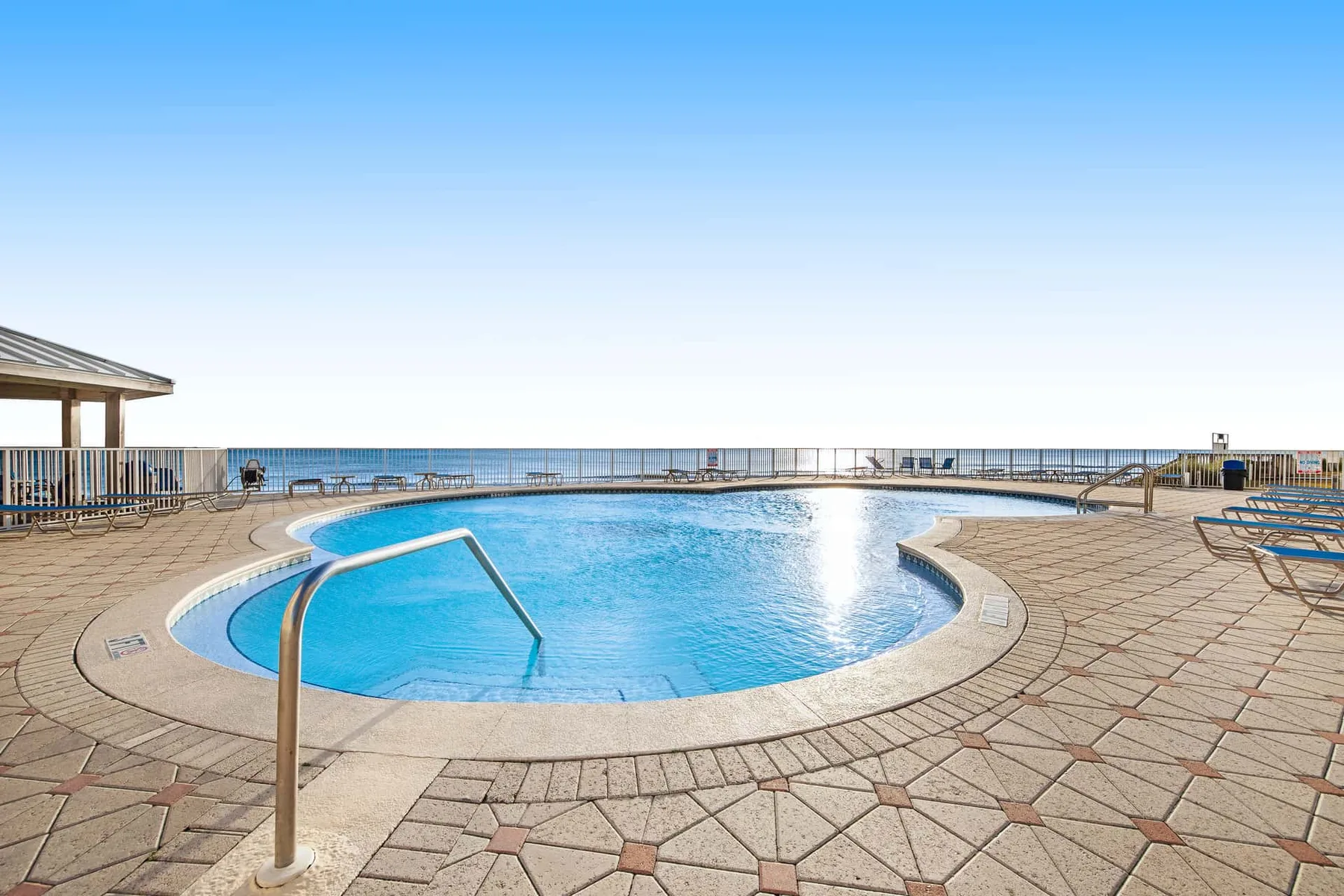A curved swimming pool sits on a paved terrace overlooking the sea.