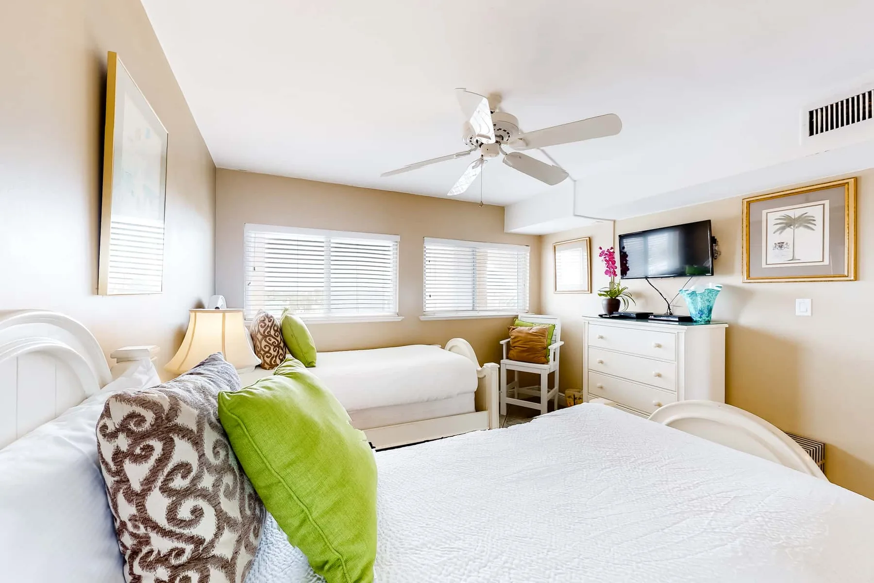 A bright bedroom with white bedding, a ceiling fan, and a television.