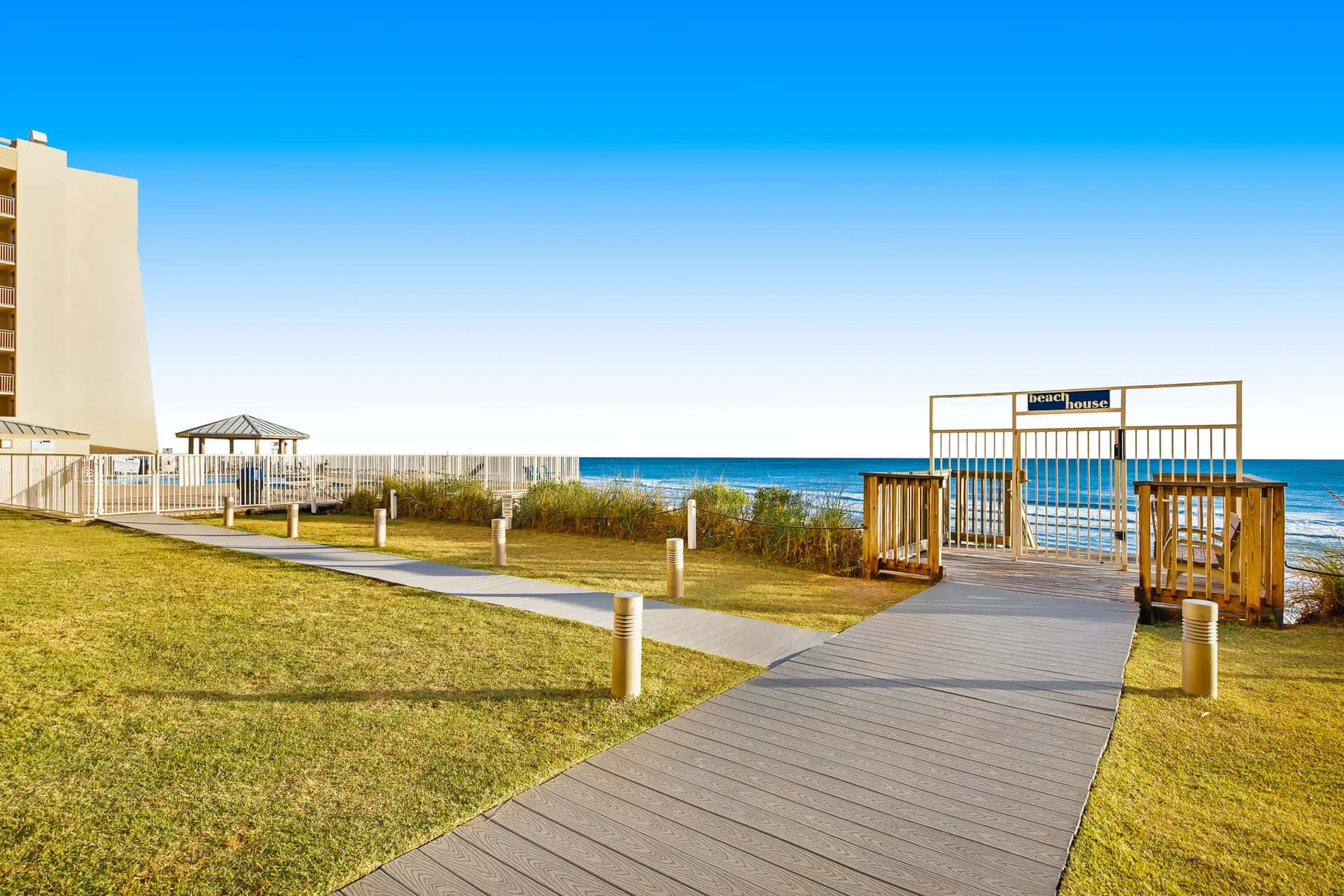 A boardwalk crosses a grassy beachfront area toward a wooden gate and the sea.
