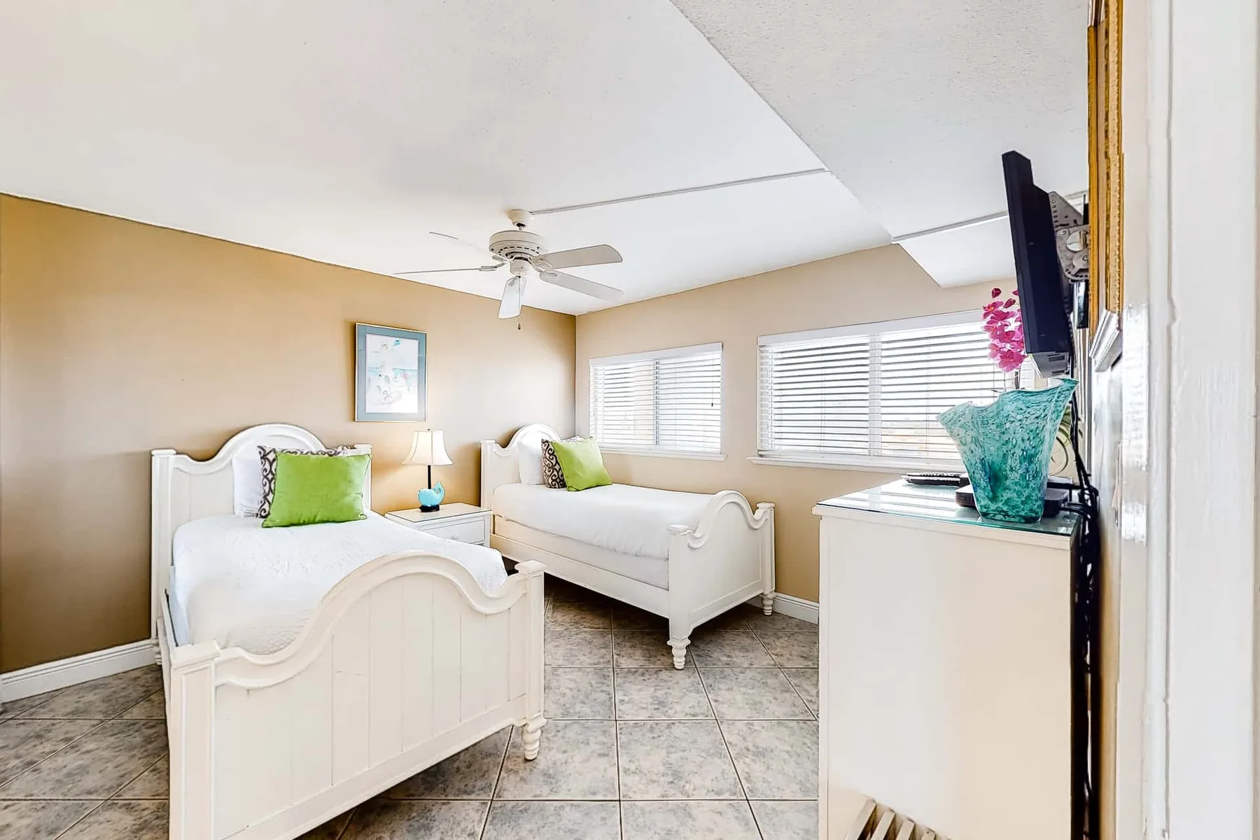 A bright bedroom with two beds and windows fitted with blinds.