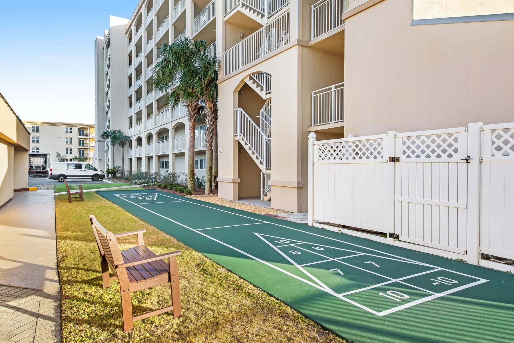 An outdoor shuffleboard court runs beside a multi-story building and a white fence.