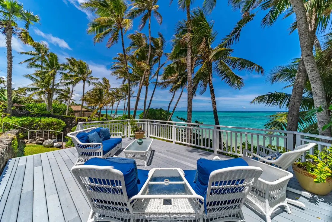 Una terraza junto al mar tiene asientos blancos y vistas de palmeras y agua turquesa.