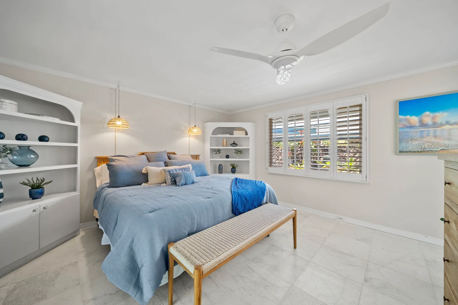 Un dormitorio luminoso con una cama azul, estanterías empotradas y ventanas con contraventanas.