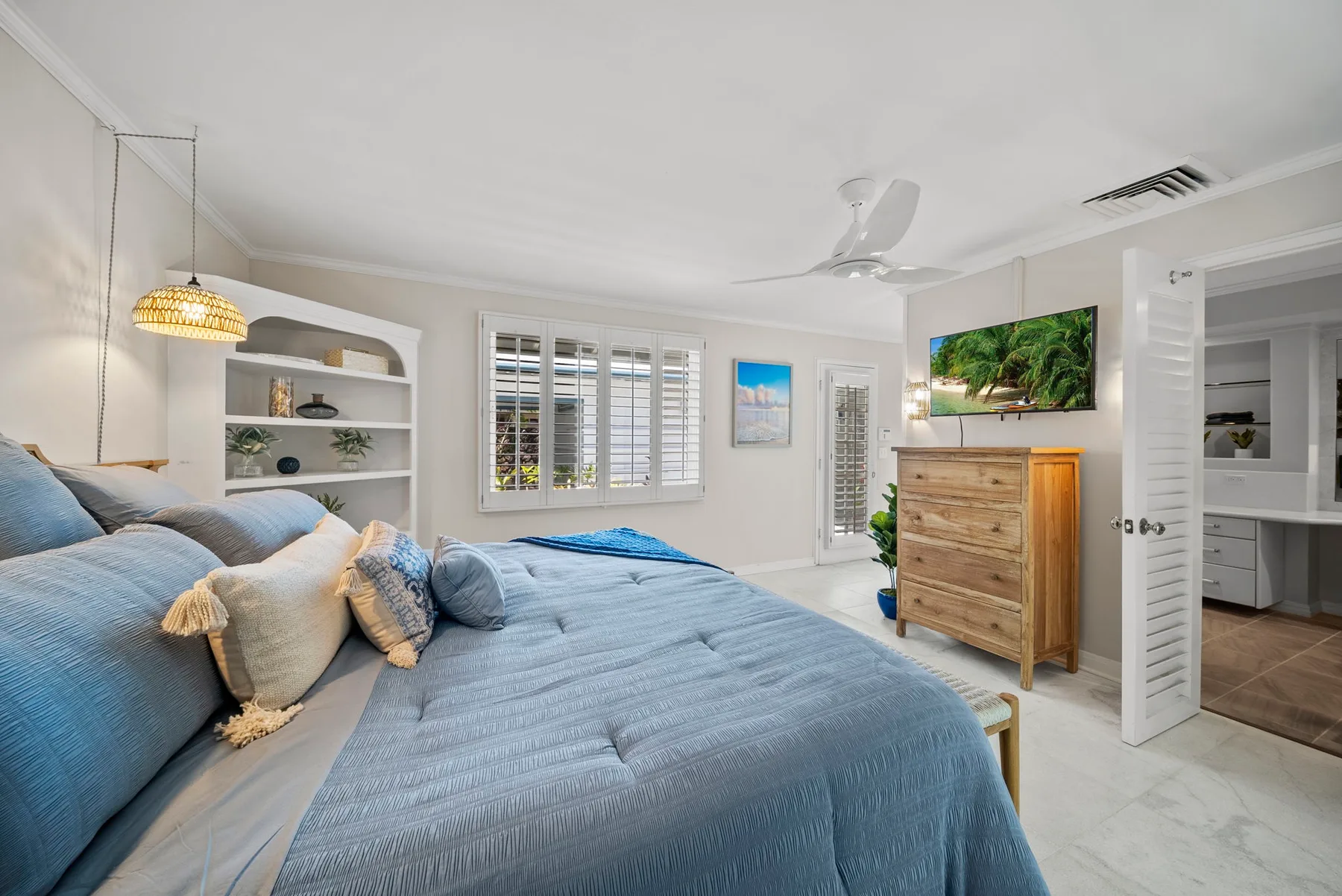 Un dormitorio luminoso con una cama bajo ventanas con persianas y una puerta abierta.