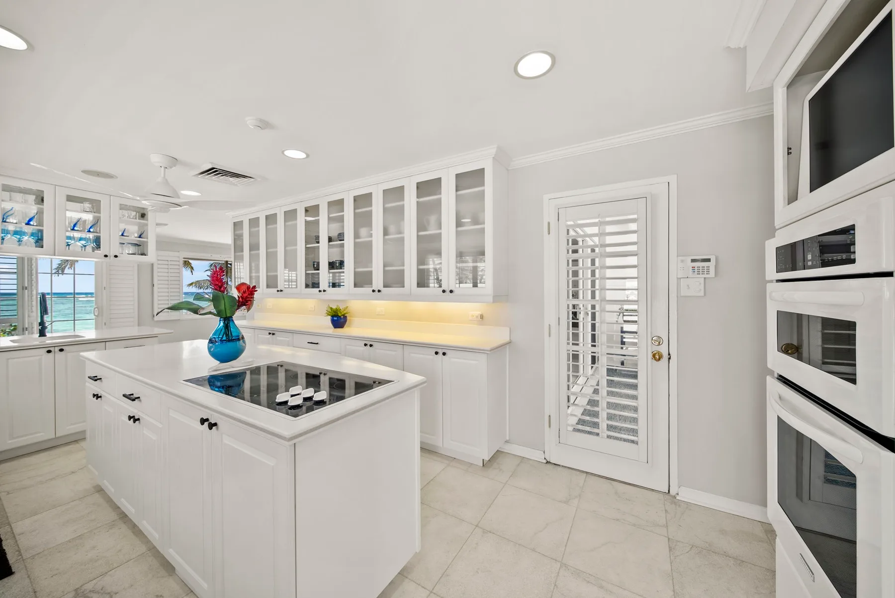 Una cocina blanca y luminosa con isla central, armarios con puertas de vidrio y electrodomésticos empotrados.