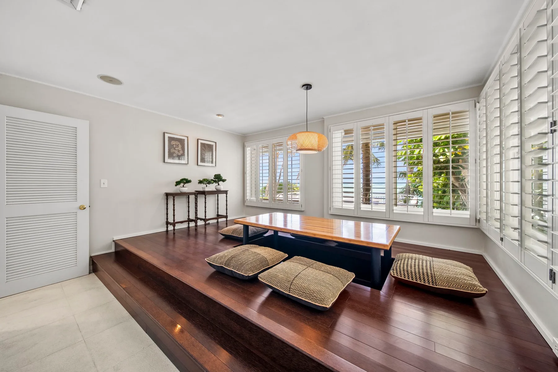 A bright sitting area with low seating around a wooden table beside shuttered windows.