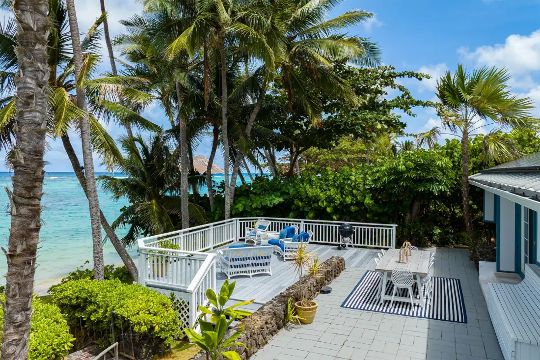 A seaside patio with outdoor seating overlooks the water beneath palm trees.
