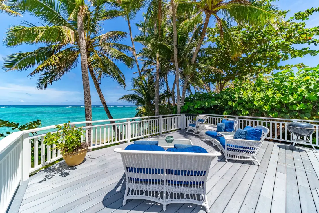 Una terraza junto al mar con asientos blancos, cojines azules, palmeras y vistas al océano.