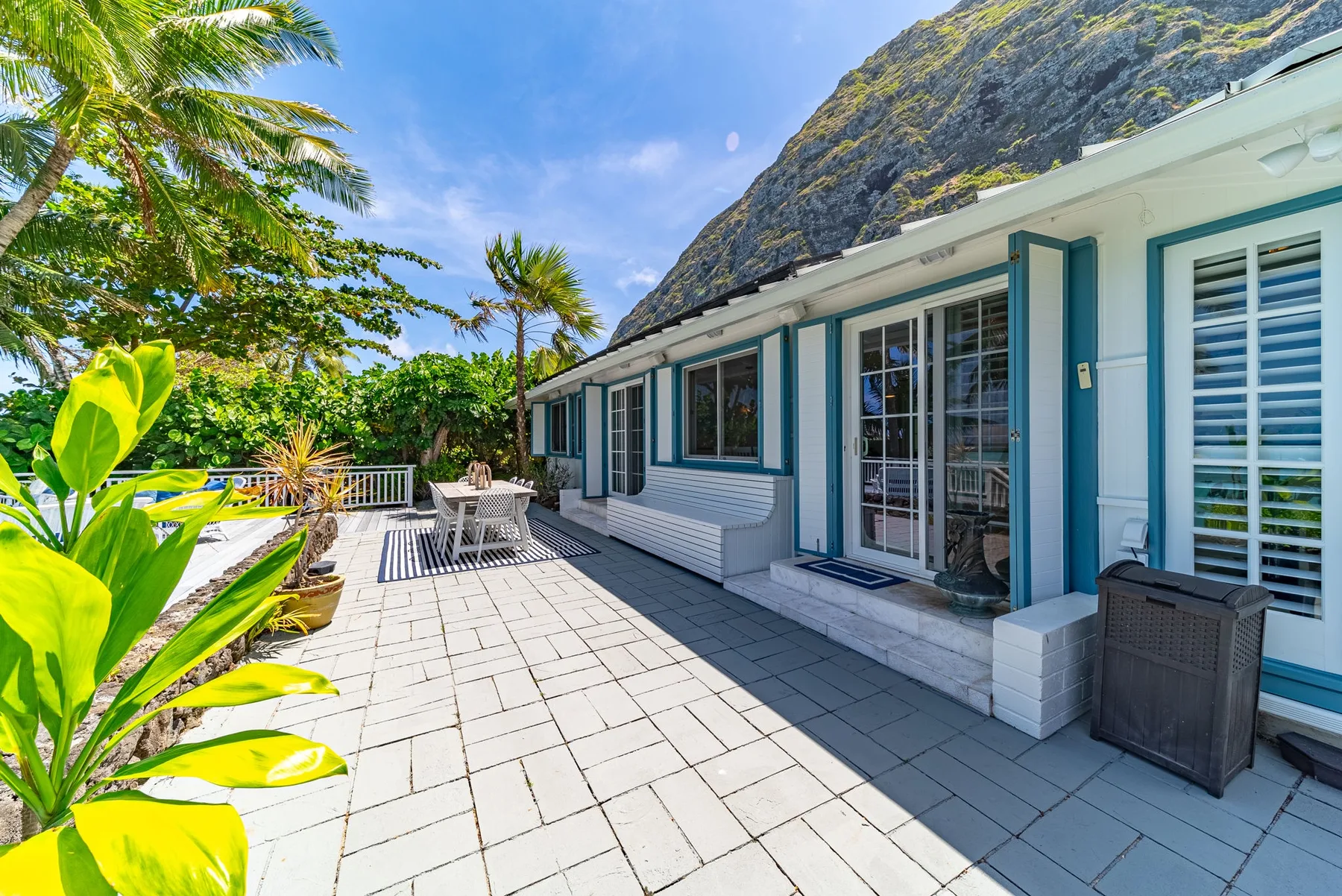 Un patio exterior embaldosado junto a una casa clara, con asientos y plantas tropicales.