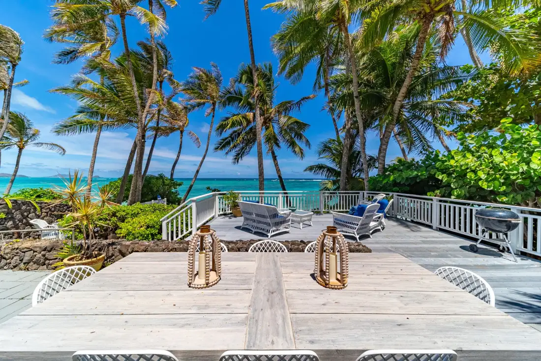An outdoor patio with a dining table and seating overlooks the ocean beneath palm trees.