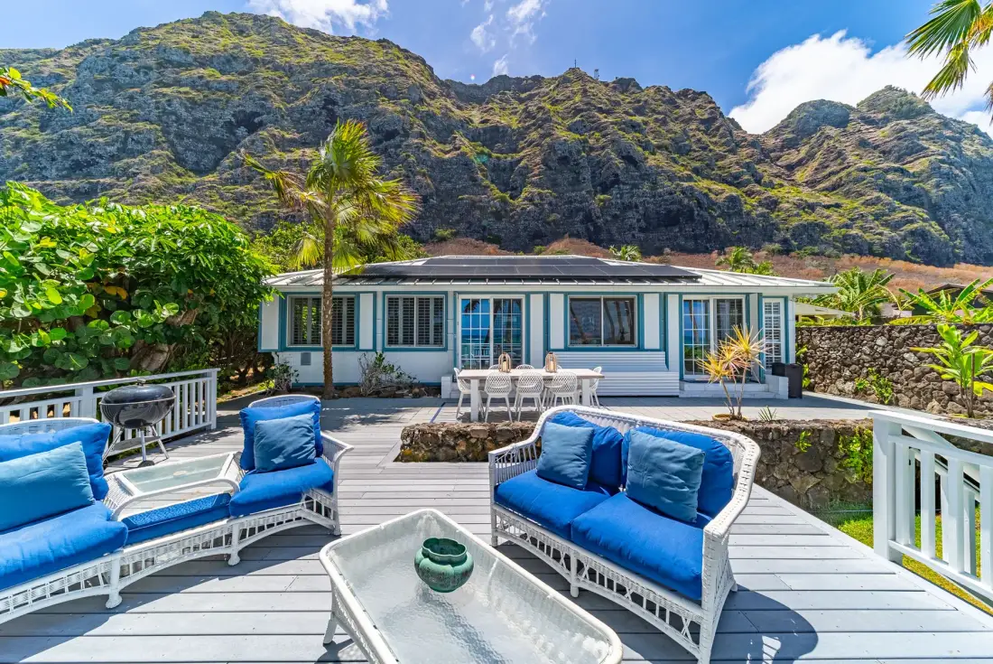 A mountain-backed house overlooks a deck with blue-cushioned outdoor seating.