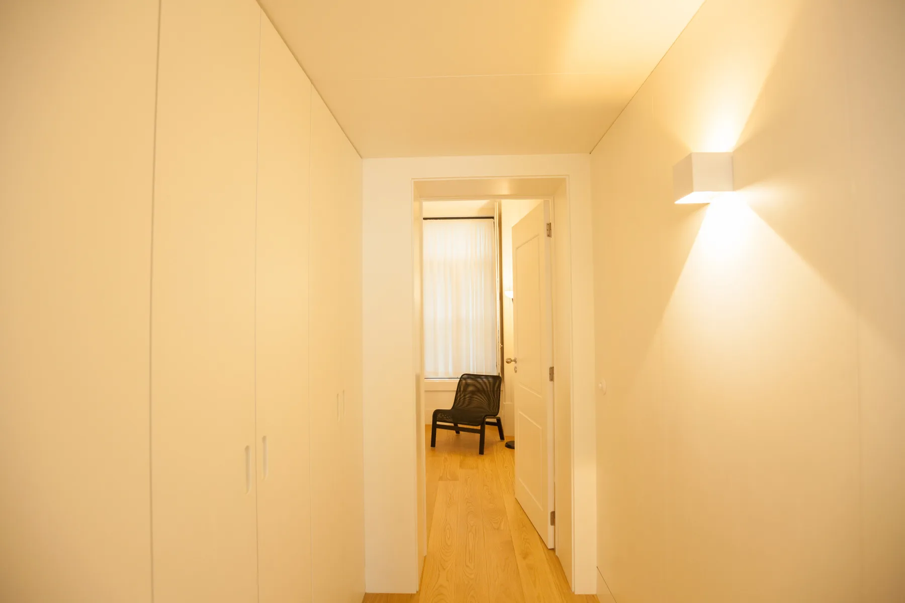 Un pasillo luminoso con armarios empotrados, una lámpara de pared y asientos al fondo.