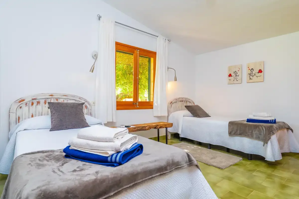 A bright bedroom with twin beds, folded towels, and a window overlooking greenery.