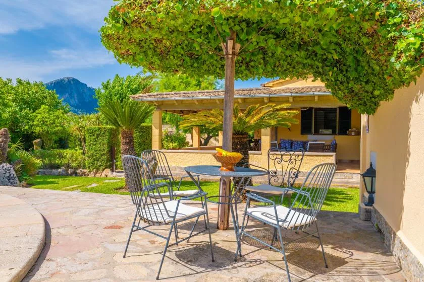 A shaded outdoor patio has metal seating and a table beneath a leafy tree.