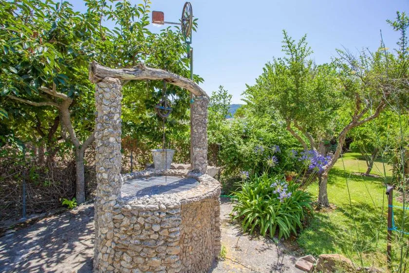 A stone well stands among trees and flowering plants in a garden.