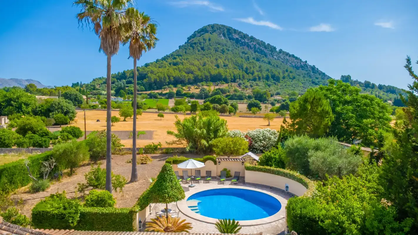 A circular swimming pool sits among gardens, with fields and a mountain in the background.