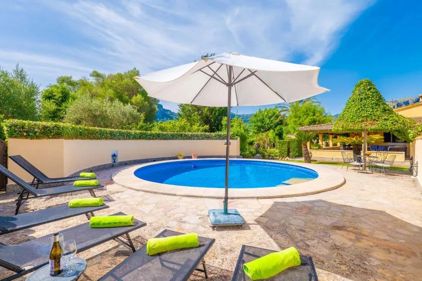 An outdoor pool is surrounded by paved decking, lounge seating, and a large umbrella.