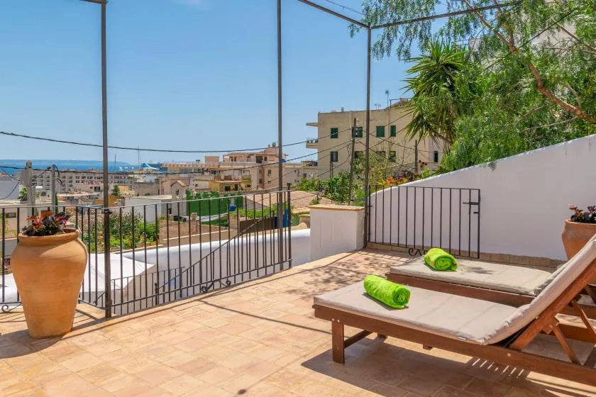 Una terraza soleada tiene asientos reclinables, plantas en macetas y vistas a edificios cercanos y al mar.