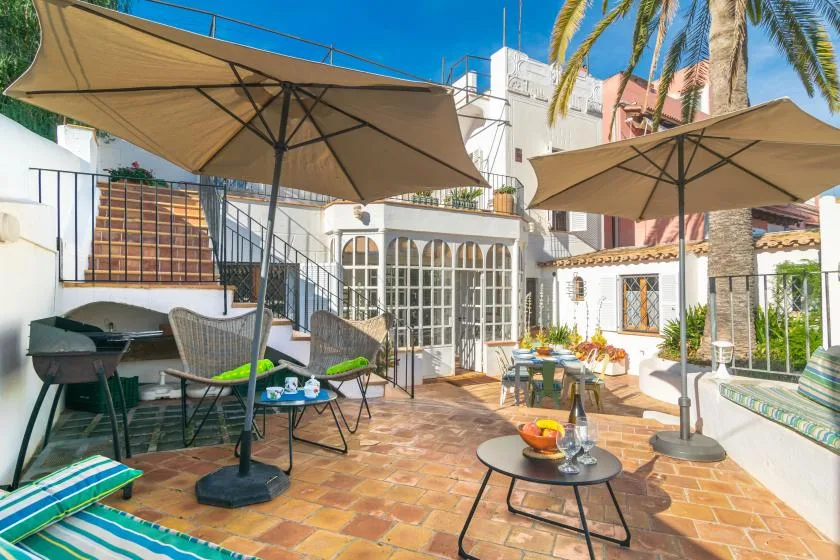 Un patio soleado con suelo de baldosas, asientos al aire libre, sombrillas grandes y plantas en macetas.