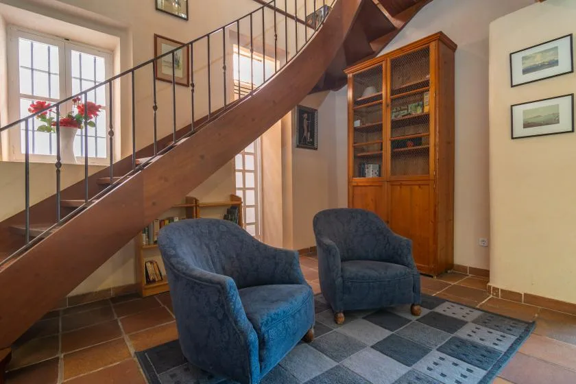 A sitting area beneath a staircase, with two blue upholstered seats and a wooden bookcase.