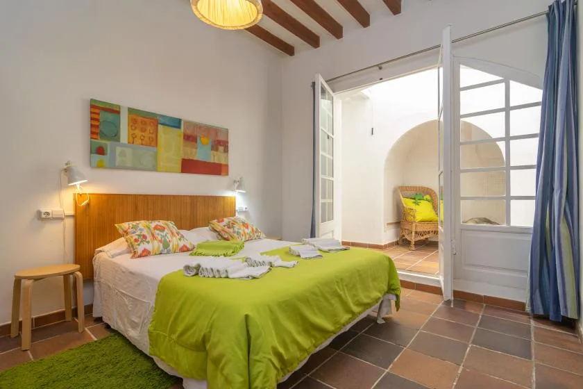 A bedroom with a bed dressed in a bright green cover beside open doors to a bright adjoining space.