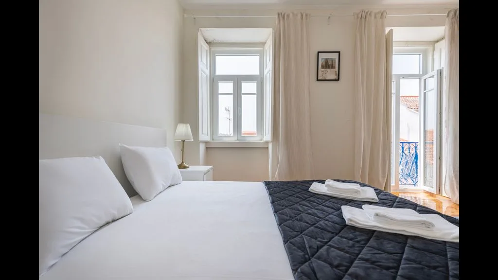 Un dormitorio luminoso con una cama junto a ventanas altas y cortinas claras.