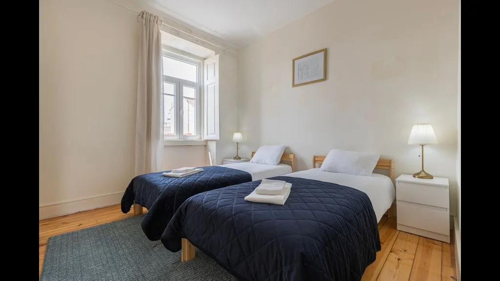 Un dormitorio luminoso tiene dos camas con colchas oscuras junto a una ventana grande.