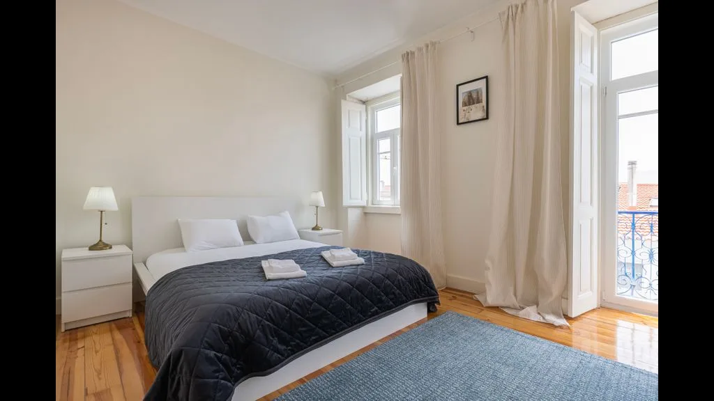 Un dormitorio con una cama, una mesita de noche y ventanas altas con vistas a un balcón.