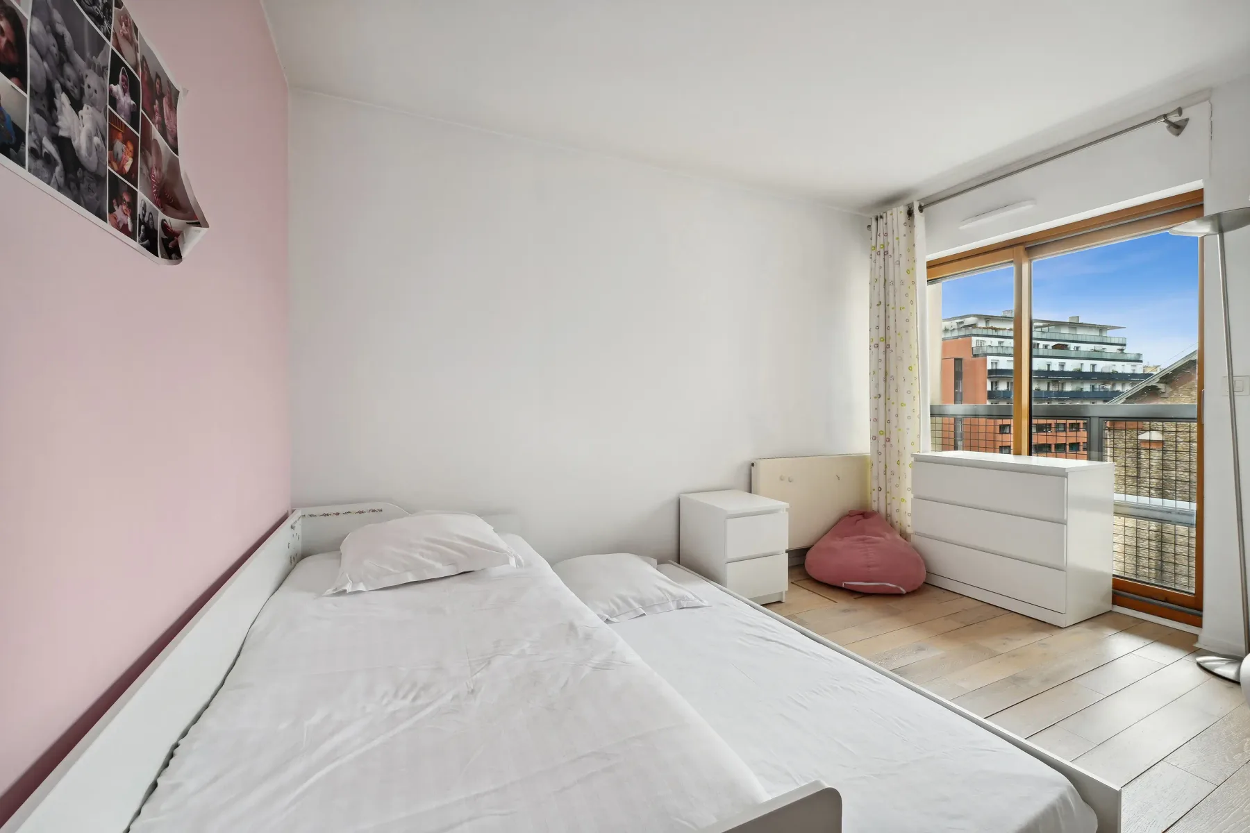 A bright bedroom with two beds and a large window overlooking nearby buildings.
