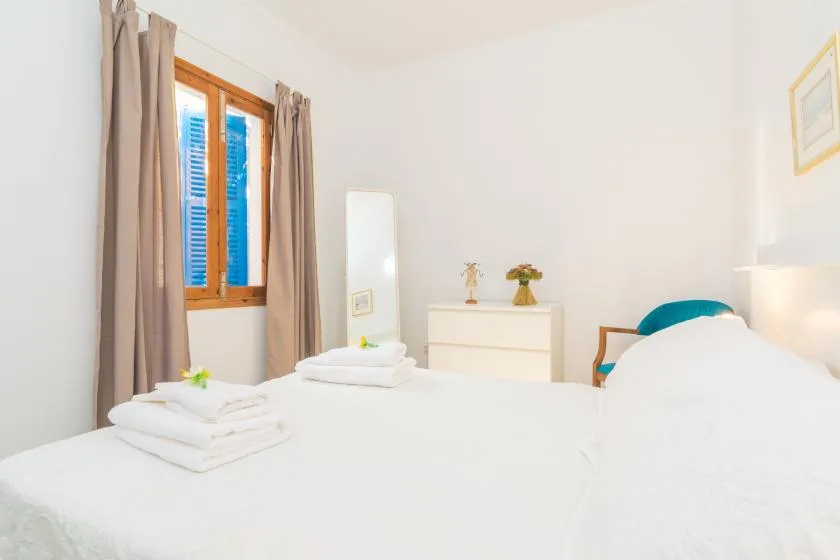 A bright bedroom with white bedding, a curtained window, and a small dresser.