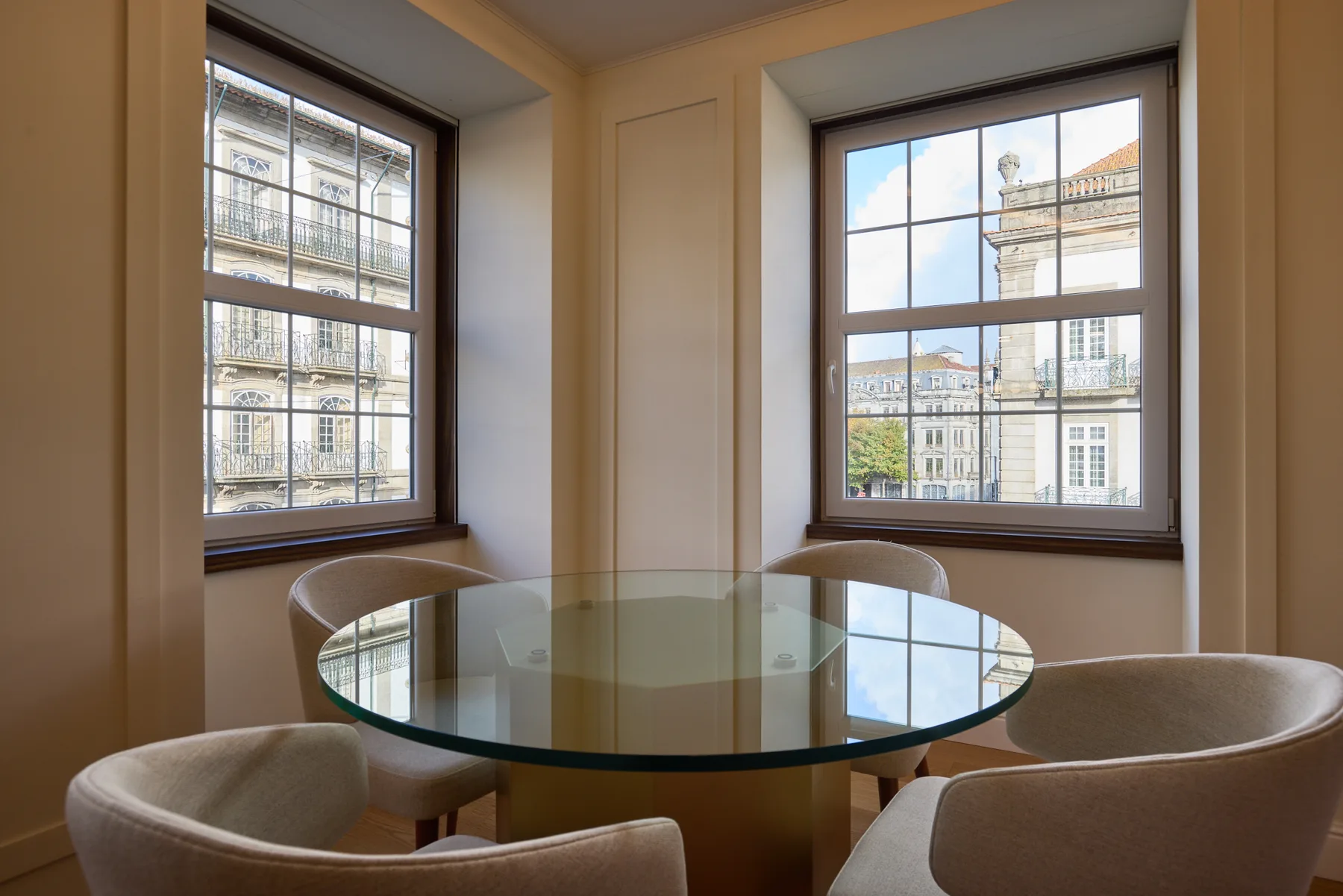 A dining area with a round glass table beside two large windows.